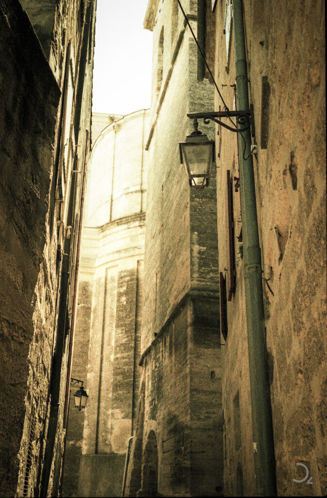Das Bild zeigt eine enge Gasse, deren hohe, verwitterte Mauern eine ruhige, nostalgische Stimmung erzeugen. Die Straßenlaterne, die an einer Hauswand hängt, verstärkt das Gefühl von Vergänglichkeit. Die warme Tönung und das weiche Licht verleihen der Szene eine zeitlose, fast geheimnisvolle Atmosphäre, als ob sie Geschichten aus der Vergangenheit in sich birgt.