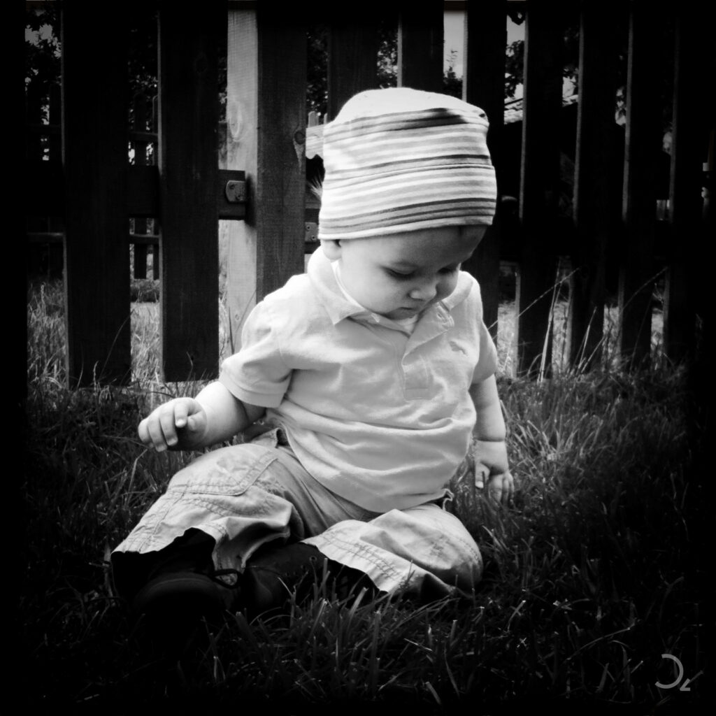 Dieses Bild im Lomografie-Stil zeigt ein Kind, das ruhig im Gras sitzt und in Gedanken versunken den Boden betrachtet. Die Schwarz-Weiß-Tonung und die leichte Vignettierung verleihen dem Bild eine nostalgische, intime Stimmung. Das Lichtspiel hebt sanft die Details im Gesicht und der Kleidung hervor, während der dunkle Holzzaun im Hintergrund Tiefe und Struktur bietet. Die Mischung aus Unschuld und Melancholie verleiht dem Bild eine besondere Ausdruckskraft.