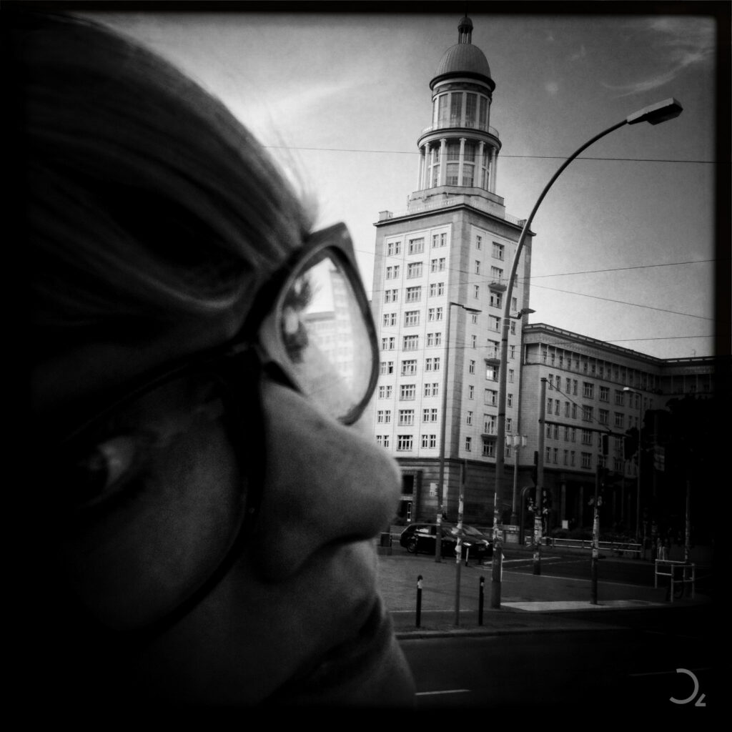 Das Bild zeigt eine Schwarzweißaufnahme mit einem markanten Gebäude im Hintergrund, vermutlich im Stil der Architektur des frühen 20. Jahrhunderts. Im Vordergrund ist eine Person mit Brille zu sehen, deren Gesicht nur teilweise sichtbar ist. Die Perspektive und das Spiel mit Nähe und Distanz schaffen eine interessante Tiefe und verleihen der Szene eine besondere Atmosphäre, die urban und zugleich persönlich wirkt.