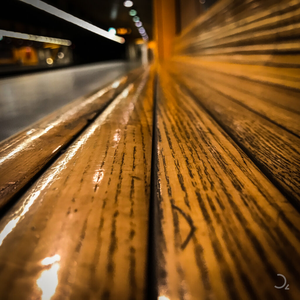 Das Bild zeigt eine Nahaufnahme einer Holzbank, die sich in die Unschärfe eines Bahnhofs erstreckt. Die glänzende Holzmaserung führt das Auge in die Tiefe, während im Hintergrund verschwommene Lichter eine warme, einladende Atmosphäre schaffen.
