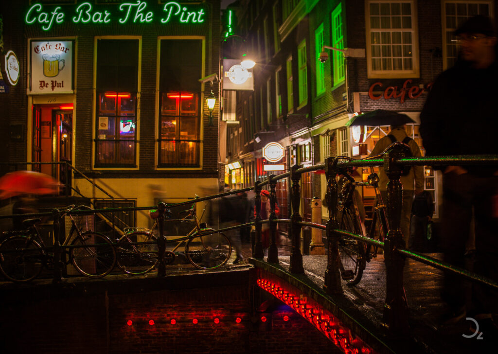 Eine nächtliche Szene an einem Kanal in Amsterdam, beleuchtet von bunten Neonlichtern und dem warmen Schein der Straßenlaternen. Im Vordergrund stehen Fahrräder entlang der Brücke, während verschwommene Silhouetten von Passanten Bewegung und Leben in das Bild bringen. Die Lichter des ‚Cafe Bar The Pint‘ spiegeln sich auf dem nassen Gehweg wider, und unter der Brücke glühen rote Lichter, die die Szenerie in ein sanftes, mystisches Leuchten hüllen.