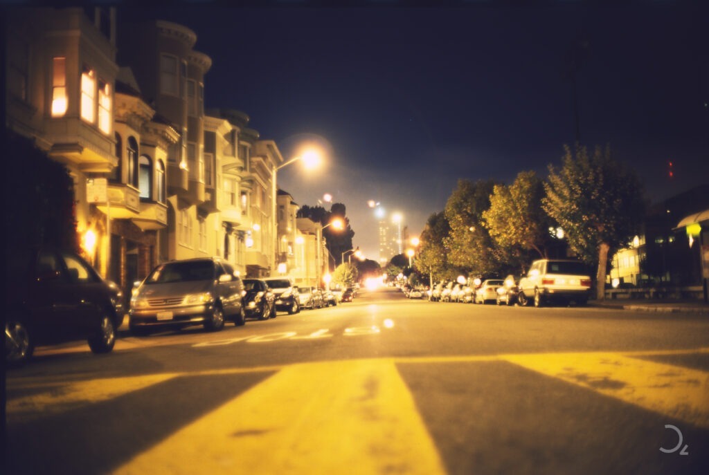 Lomography, die Magie des Zufalls. Stimmungsvolle Nachtaufnahme einer ruhigen, von Bäumen und historischen Gebäuden gesäumten Straße, beleuchtet von warmem Straßenlicht. Autos parken entlang der Straße, und in der Ferne sind verschwommene Lichter der Skyline San Franciscos zu sehen.