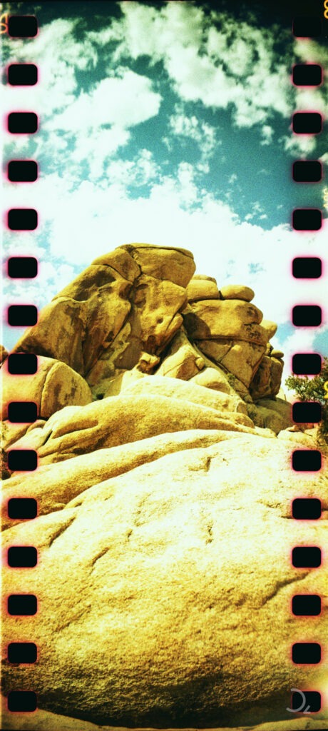 Analoge Aufnahme von großen Felsformationen unter einem teilweise bewölkten Himmel, eingerahmt von den typischen perforierten Filmstreifenrändern. Die warmen Farben und die Struktur der Felsen verleihen der Szene eine natürliche und raue Ästhetik.