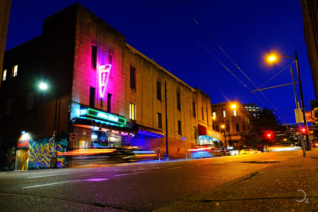 Street Photography, die Kunst des Augenblicks. Die Nacht war in die Stadt eingezogen und die Lichter erwachten zum Leben. Neonlichter blitzen an den Wänden der alten Gebäude und verliehen der Straße ein pulsierendes Flair. Autos fuhren vorbei, ihre Scheinwerfer hinterließen flüchtige Lichtspuren auf dem Asphalt. Ein Gefühl von Energie und Möglichkeiten lag in der Luft, während Menschen in der Dämmerung ihren Weg suchten. Die Stadt schien voller Geschichten, die darauf warteten, erzählt zu werden.