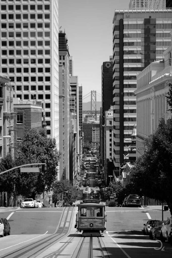 Eine ikonische Szene von San Francisco: Die historische Cable Car fährt die steilen Hügel hinauf, während die massiven Gebäude beidseitig die Straße säumen. In der Ferne thront die Bay Bridge, die sich wie ein stummer Wächter über die Bucht spannt. Der Anblick verbindet Nostalgie mit Moderne und erzählt die Geschichte einer Stadt, die sich zwischen Vergangenheit und Gegenwart bewegt. Der Straßenlärm und das leise Rattern der Schienen scheinen im Kontrast zur majestätischen Stille der Brücke zu stehen. Ein Bild des pulsierenden Lebens und der bewegten Geschichte San Franciscos.