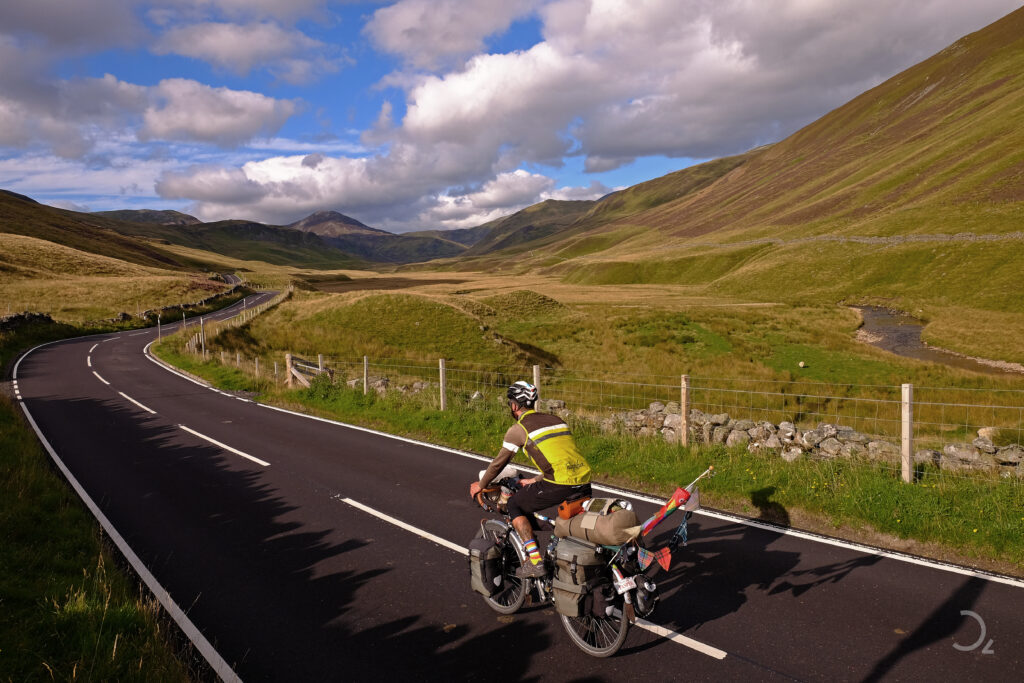 Dieses Bild zeigt einen Radfahrer, der auf einer ruhigen, kurvigen Straße in einer malerischen schottischen Landschaft unterwegs ist. Die üppigen, sanften Hügel und das klare, sonnige Wetter schaffen eine idyllische Szene. Der Radfahrer, beladen mit Taschen und Ausrüstung, scheint die Weite und Schönheit der Umgebung zu genießen. Schatten von Wolken fallen auf die Hügel und betonen die sanften Erhebungen der Landschaft. Ein kleines Flussbett schlängelt sich durch das Tal, das durch die unberührte Natur und das Spiel von Licht und Schatten sehr lebendig wirkt.