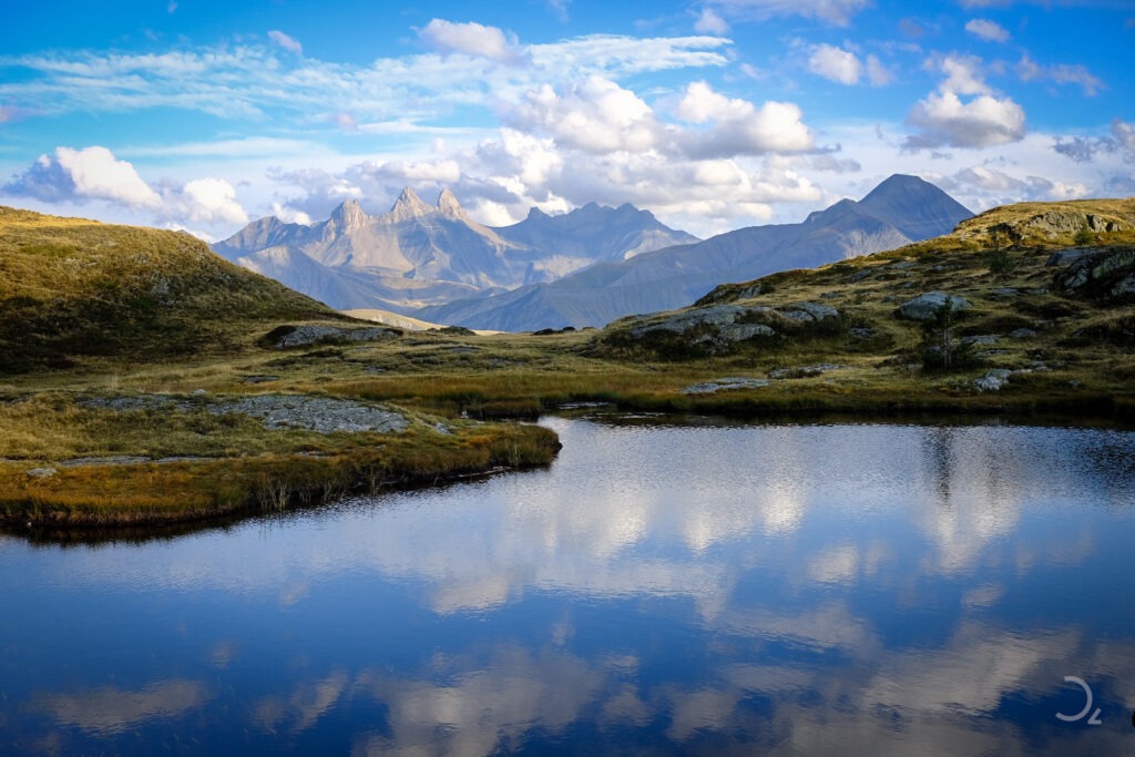 Landscape Photography - Inmitten einer beeindruckenden Berglandschaft spiegelt sich der klare Himmel im ruhigen Wasser eines kleinen Teichs. Die sanften Hügel und schroffen Gipfel der Umgebung werden von einem warmen Licht erleuchtet, während die Wolken in der Ferne majestätisch vorbeiziehen. Die friedliche Szenerie lädt zum Verweilen ein und lässt den Betrachter die Schönheit der Natur in vollen Zügen genießen.
